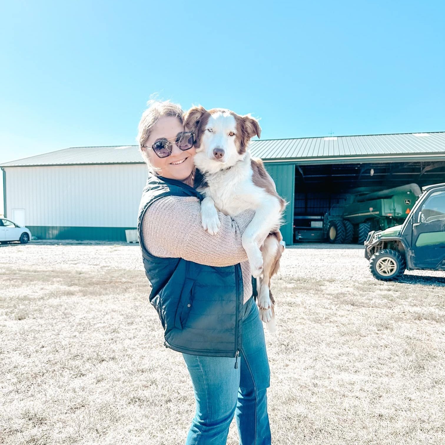 Woman holding a dog outdoors on a sunny day, wearing sunglasses and a cozy sweater, with a farm setting in the background featuring a green barn and vehicles.