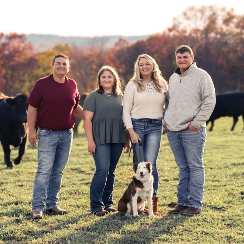 Family portrait in a scenic farm setting, featuring four smiling individuals and a dog, with cows grazing in the background and autumn foliage. Ideal for showcasing rural lifestyle and family values.