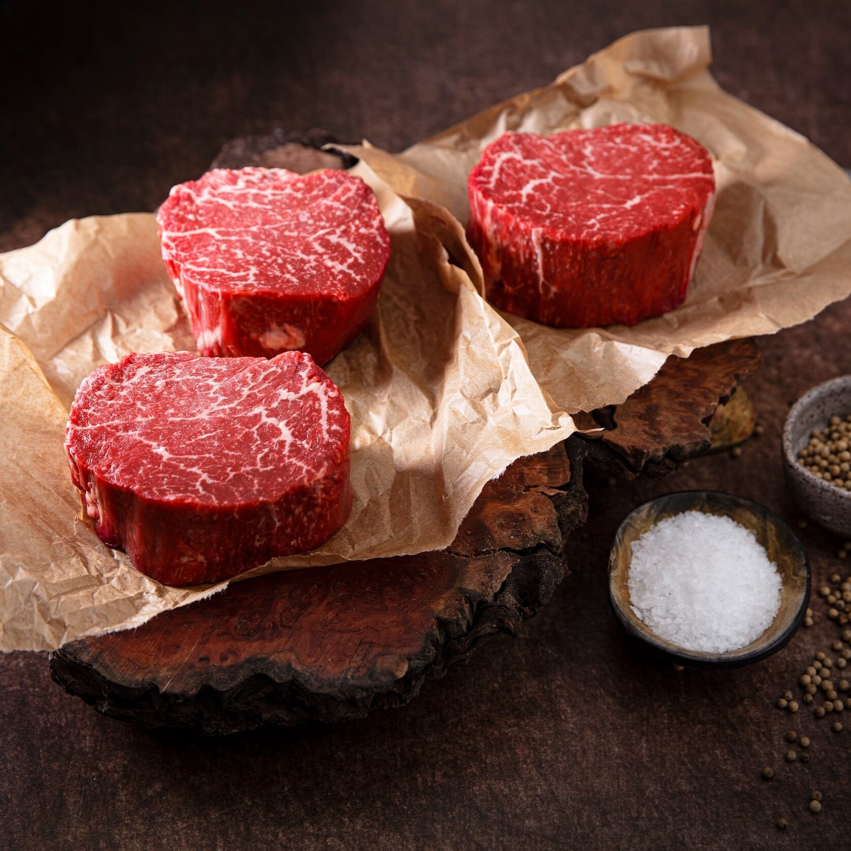 Three cuts of raw marbled beef steak on crumpled parchment paper, displayed on a rustic wooden board, accompanied by bowls of salt and peppercorns.